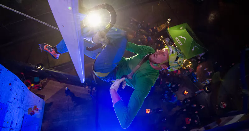 Een klimmer met een helm, handschoenen en een groene jas beklimt een klimwand in een indoorhal met behulp van een ijsbijl, terwijl er achter hem een fel licht schijnt en er beneden mensen staan te kijken.